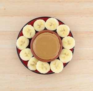 Top view of sliced bananas on a red plate in a circle around a bowl of peanut butter on a wood table.